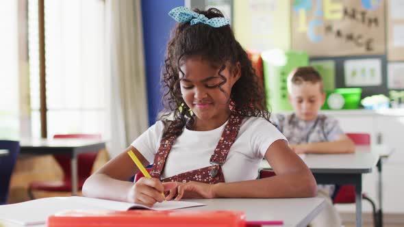 Portrait of happy mixed race schoolgirl sitting at classroom, making notes, looking at camera