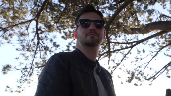 Young bearded man with sunglasses and leather jacket stands in sunshine under a tree. Panning shot