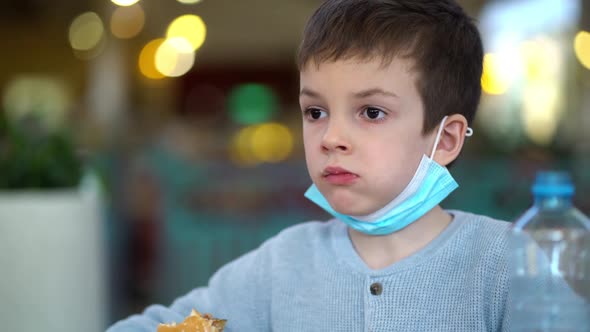 Boy with Medical Mask Eats a Burger with Cutlet Sitting in Restaurant