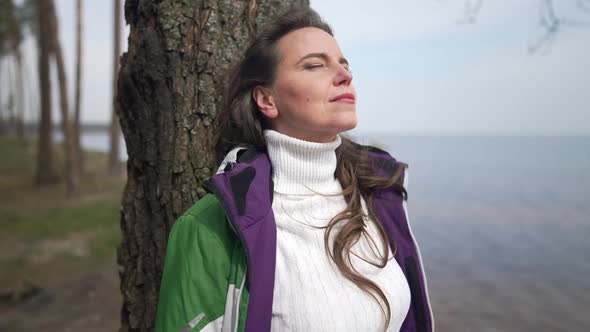 Relaxed Caucasian Woman Standing at Tree Trunk on River Bank Breathing Fresh Air in Slow Motion