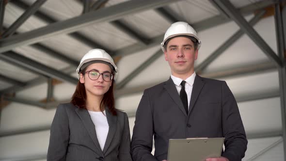 Young Man and Woman in Helmets at a Construction Site. Businessmen in Suits Are Looking at the