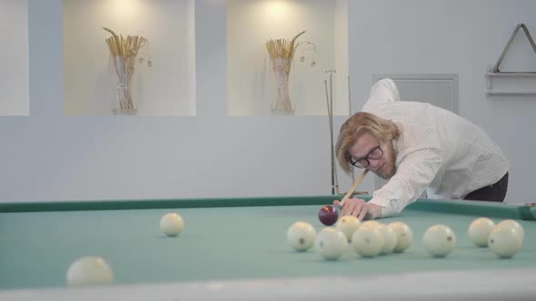 Blond Bearded Man Playing Pool, Billiards in Light Room. Confident Player Hits the Ball with a Cue