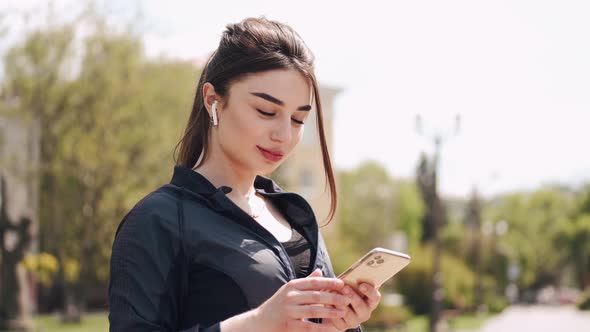 Beautiful Athletic Woman in Earpods and Sportswear Is Standing Outside with a Smartphone