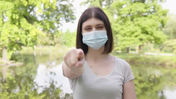 A Young Caucasian Woman in a Face Mask Points at the Camera and Nods in a Park