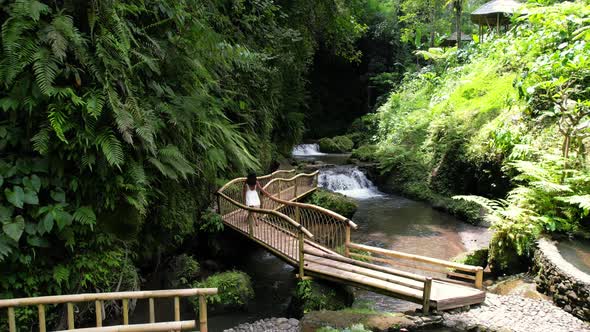 Woman in white dress walks on bamboo bridge towards waterfall river in jungle, aerial