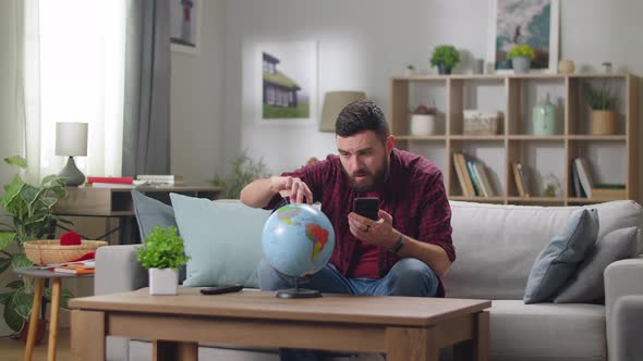 Young Handsome Guy Choosing a Country to Travel on a Globe While Sitting on a Sofa in the Living