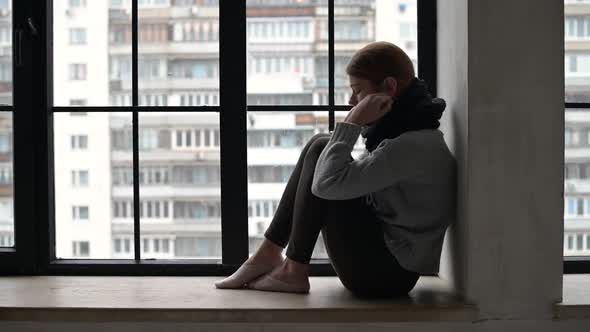 Young woman is sad sitting on the windowsill of city apartment