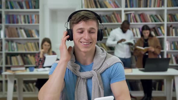 Young Man in Casual Clothes Sitting in the Library and Listening Music in Earphones