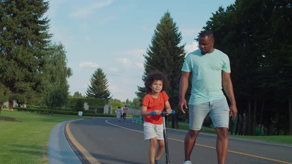 Joyful Little Son Riding Push Scooter in Park