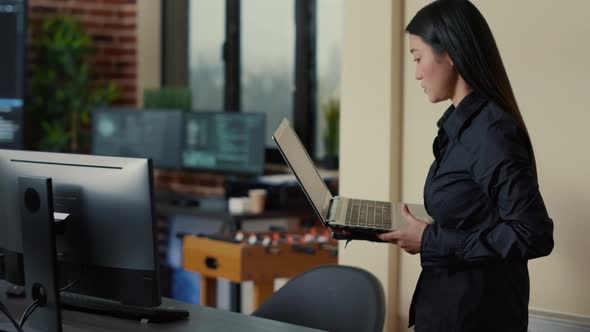 Asian Software Developer Coming at Desk and Sitting Down Holding Laptop with Coding Interface