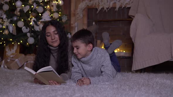 Mom Reads a Book To Her Son and Kisses Him Lying on the Floor Near the Christmas Tree