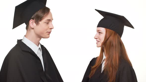 Ginger Caucasian Graduate Female Talking with Male in Black Robe and Square Academical Cap on White