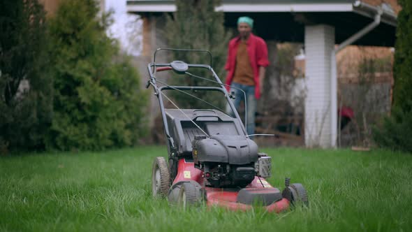 Lawn Mower in Backyard Garden with African American Young Man Walking to Equipment From Background