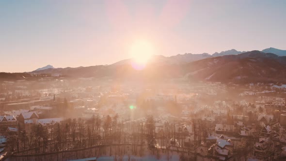 Sun Shining Behind Mountains Against A Town With Small Houses Woods In Foreground