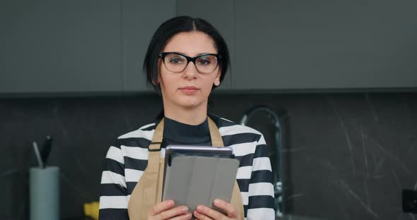 Close Up Portrait of Attractive Busy Concentrated Woman in Kitchen Apron Holding Tablet Device