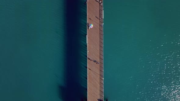 overhead shot over a long wooden pier above blue seawater, tourists strolling along