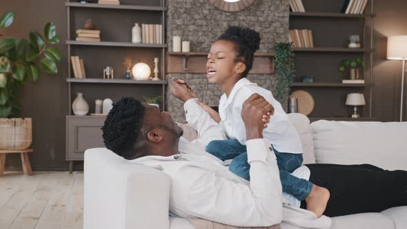 African Father Lying on Sofa at Home with Daughter Playing Fight Game Having Fun Spending Free Time