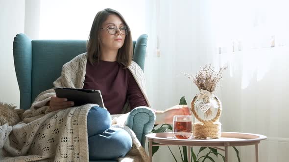 Young Woman in Eyeglasses Using Digital Tablet Drinking Tea Sitting in Armchair at Home