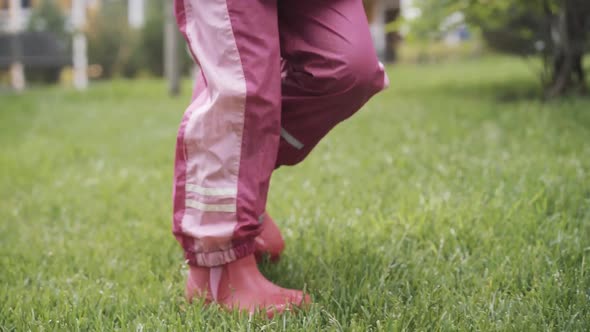 Feet of Little Girl in Pink Rubber Boots and Waterproof Pants Walking on Wet Green Grass Outdoors