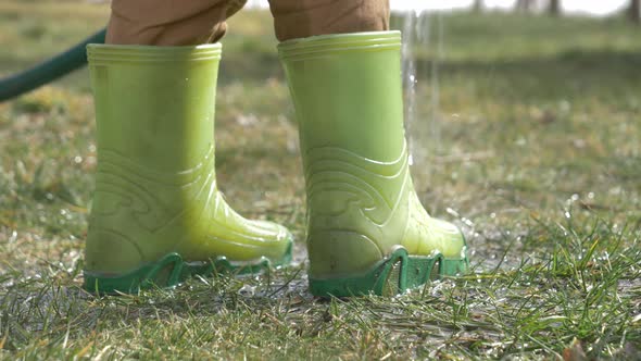 kid in green rubber boots play with water in tube in a garden alt