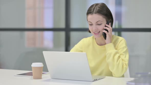Young Woman Talking on Smartphone While Using Laptop in Office