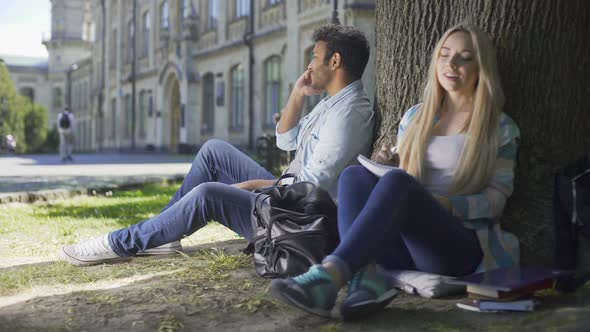 Young Male and Female Sitting Under Tree, Guy Talking on Phone, Girl Writing