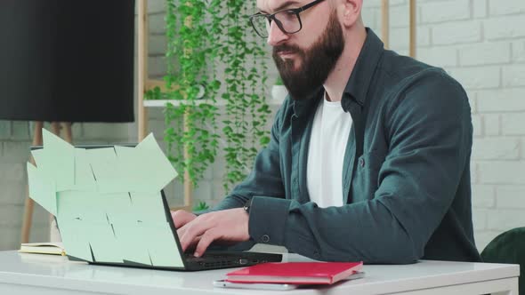 Bearded Man Types at the Laptop and Write Notice on the Paper Notebook
