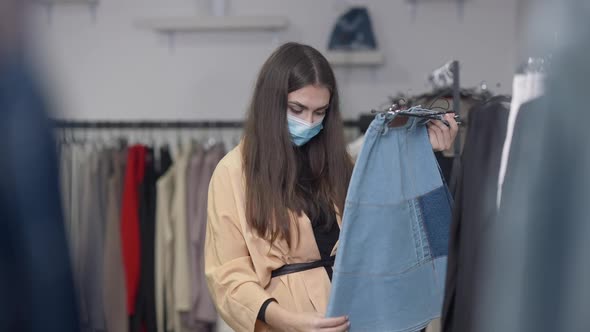 Portrait of Concentrated Woman in Coronavirus Face Mask Checking Jeans Skirt in Shop on Black Friday