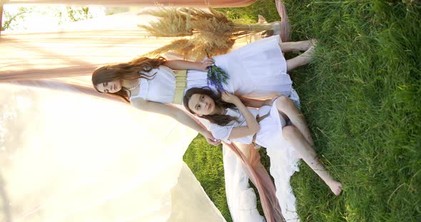 Two Girl Teenagers in White Dresses with Flowers Bouquet Posing on Chifon Background in a Field at