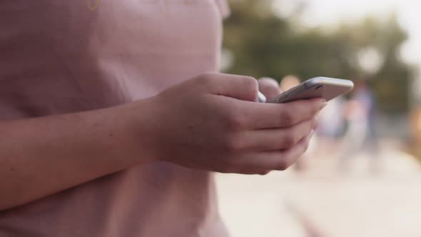Woman in park is typing message on smartphone holding it with both hands, closeup, front view.