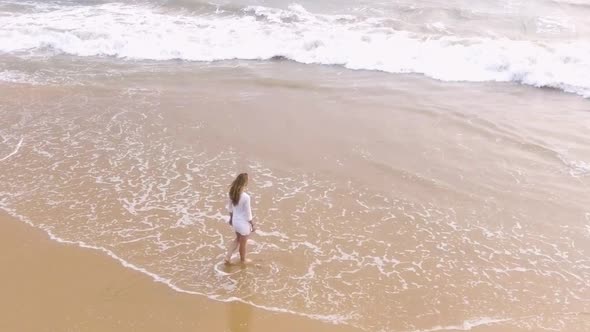 A Young Girl in a Short White Dress Is Walking Along the Sandy Beach in the Tropics, Near the Ocean