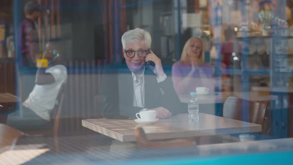 View Through Window of Senior Businessman Talking on Phone and Drinking Coffee in Modern Cafe