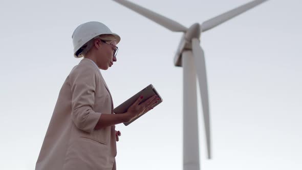 A Girl Engineer in a White Hard Hat Looks at a Digital Tablet and Holds Out Her Hand with a Battery