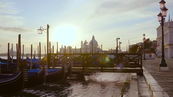 Long Gondolas Moored To Lagoon Pier Against Morning Sunlight