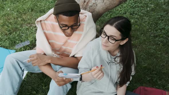 Couple of International Students Working on Homework Together Sitting on the Lawn Near the Tree