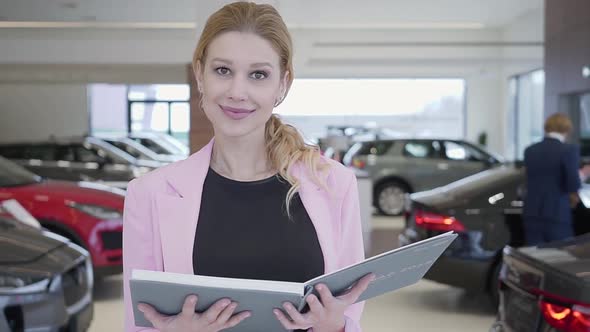 Portrait of Pleasant Woman in Pink Jacket Walking Toward Camera Holding Big Book