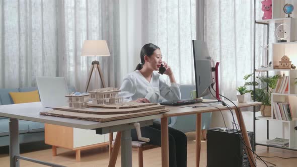 Asian Woman Engineer With The House Model Talking On Smartphone While Working On A Desktop At Home