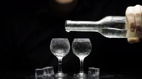 Barman Pour Frozen Vodka From Bottle Into Two Glasses with Ice. Black Background