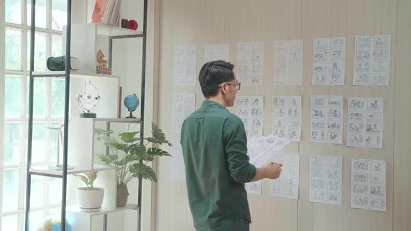 Young Creative Asian Designer Looks At The Storyboard Sketches Covering His Wall