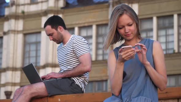 Man Using Laptop and Woman Using Smartphone Sitting on a Bench Outdoors