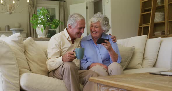 Caucasian senior couple smiling while using smartphone together sitting on the couch at home