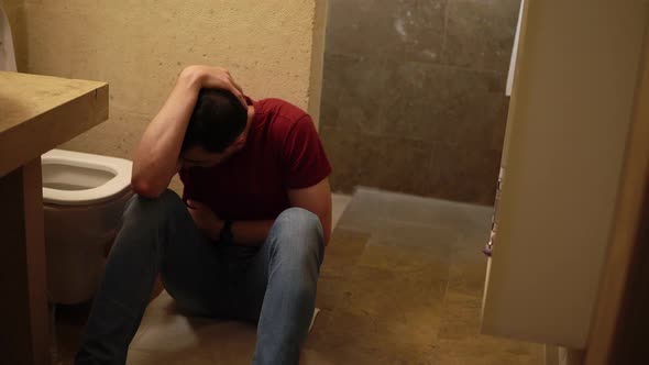 Young Sick Guy is Sitting on the Floor of Bathroom Next to Toilet and Holding His Belly