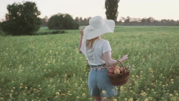 A Young Read Head Woman with Hat Holding a Woven Basket of Peaches and Walking on a Grass Path