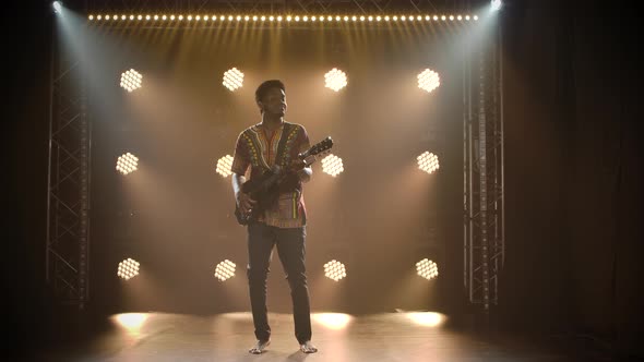 Musician in Ethnic African Costume Plays the Guitar in a Dark Studio. Black Barefoot Man Closed His