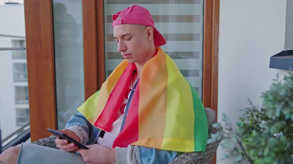 Homosexual Man Covered with Rainbow Flag Using Tablet While Sitting on the Terrace