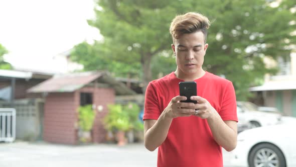 Happy Young Asian Man Smiling While Using Phone Outdoors