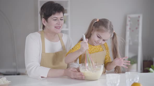 Little Pretty Girl Sitting with Grandmother in Kitchen and Beating Eggs with Flour. Cute Caucasian