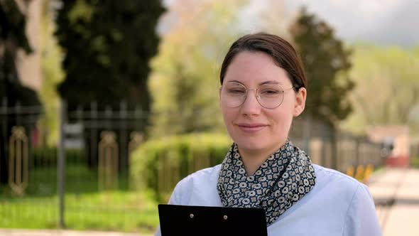 Girl Student with Glasses is Walking Down the Street with a Folder in Her Hands