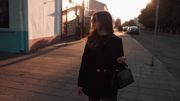 A Gorgeous Young Girl in Black Clothes and Sunglasses Stands on a City Street at Sunset and