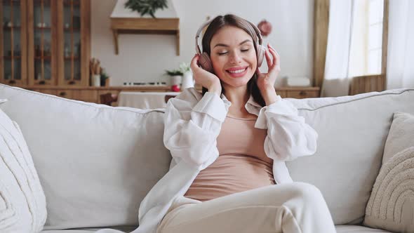 Positive Pregnant Woman Listens to Music Sitting on Sofa